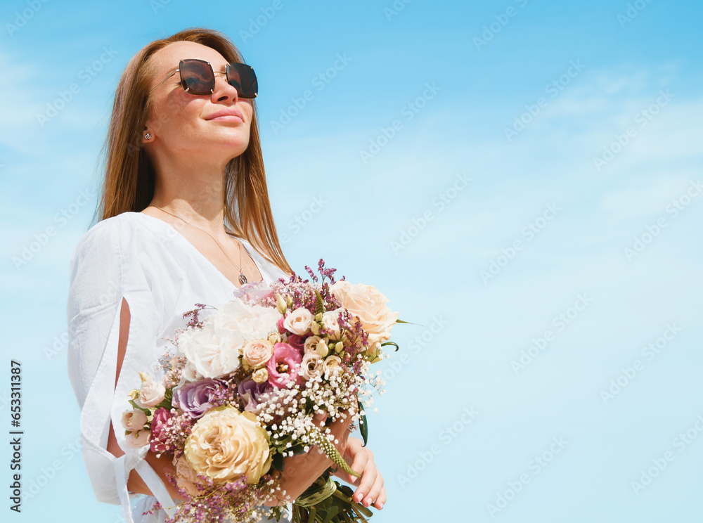 Joyful Young Woman Holding a Bouquet of Flowers on a Beach with Blue Sky Background
