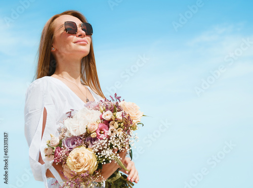 Obraz na płótnie Joyful Young Woman Holding a Bouquet of Flowers on a Beach with Blue Sky Background w salonie