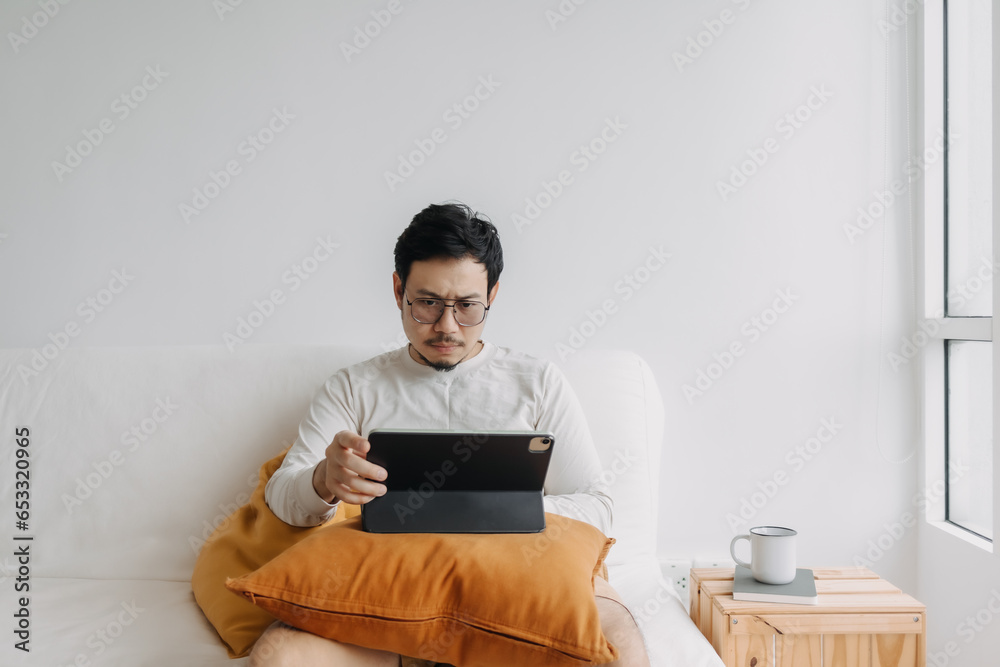 Asian man working using tablet, sitting on sofa at apartment, thinking something seriously, wearing sweater in cold day. Asian man working using tablet, sitting on sofa at apartment, thinking something seriously, wearing sweater in cold day.