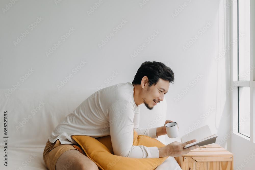 Happy Asian man enjoy reading the book and drinking coffee tea, sitting on sofa at apartment in cold weather. Happy Asian man enjoy reading the book and drinking coffee tea, sitting on sofa at apartment in cold weather.