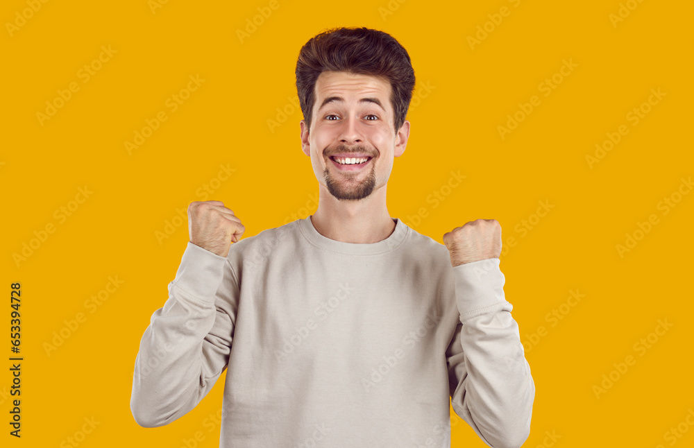 Yes, great success. Portrait of a happy, excited young man in a sweatshirt standing isolated on an orange yellow background, looking at the camera, clenching fists, doing a yes gesture and smiling Yes, great success. Portrait of a happy, excited young man in a sweatshirt standing isolated on an orange yellow background, looking at the camera, clenching fists, doing a yes gesture and smiling