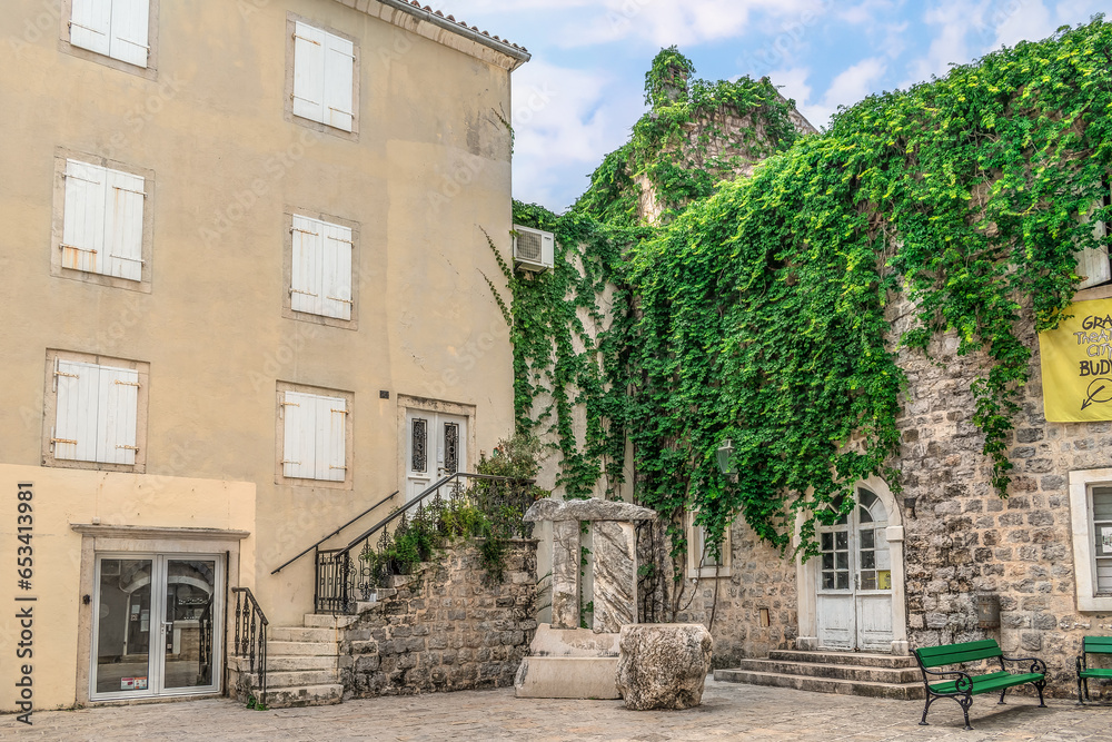Budva, Montenegro - June 17, 2021: The Poets Square in the Old Town of Budva. Stone table and chair on a city street against the backdrop of ancient medieval buildings overgrown with five-leaved ivy