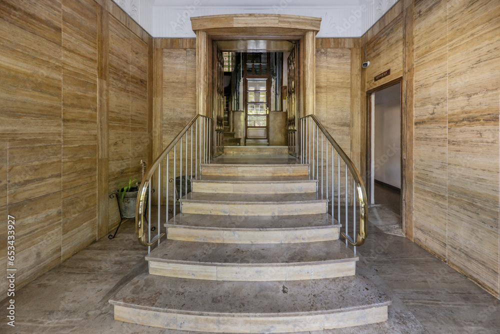 Access portal to the building with brown marble floors on the walls, stairs with golden metal railing and vintage elevator Access portal to the building with brown marble floors on the walls, stairs with golden metal railing and vintage elevator