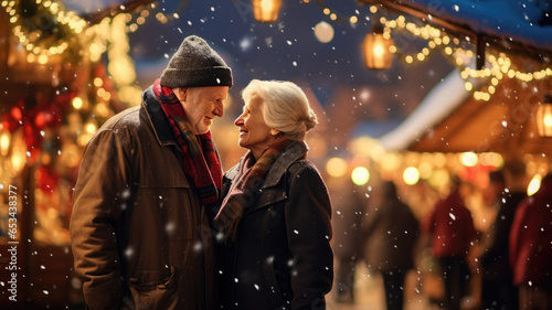 A couple of retired people in love are standing at a Christmas market on a snowy evening and smiling.