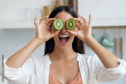 Funny young woman posing with kiwi slices in the kitchen at home.
