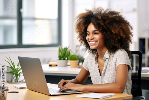 African woman with an afro, smiling and looking into her laptop computer on her desk in a clean white office
