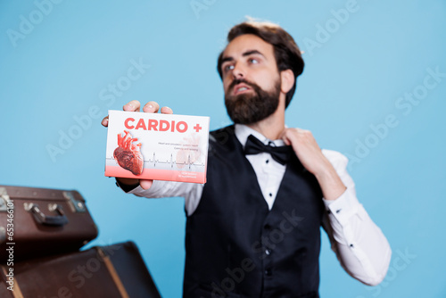 Hotel concierge presents cardiac pills in front of studio camera, recommending medication for heart disease or problems. Young man doorkeeper shows box of medicaments treatment.