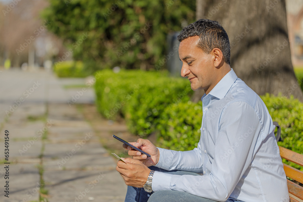 Fototapeta premium Young Latino man making a purchase with a credit card on his mobile phone sitting on a bench in a public park.