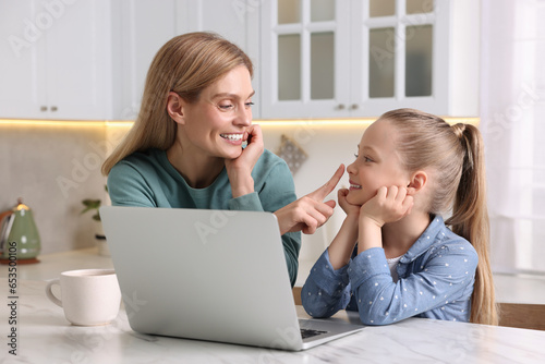 Happy woman and her daughter with laptop at white table indoors