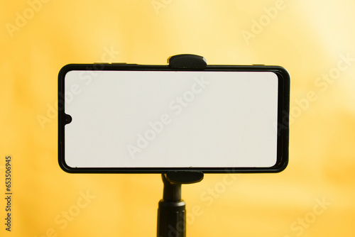 Landscape phone with white screen fixed to tripod on yellow background, for mockup design.