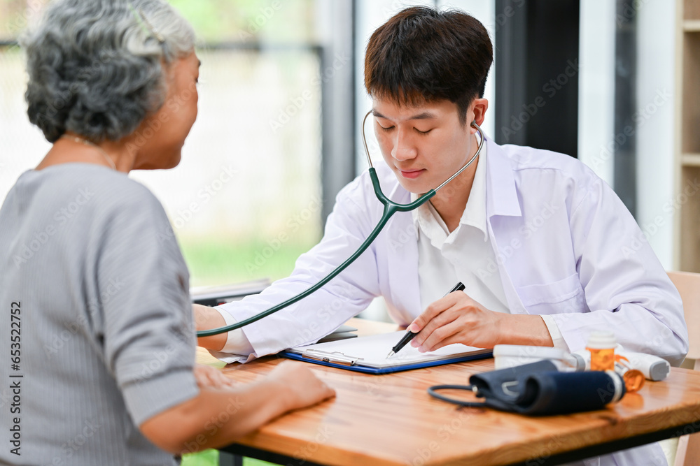 A professional Asian male doctor is listening to a patient's heartbeat with a stethoscope.