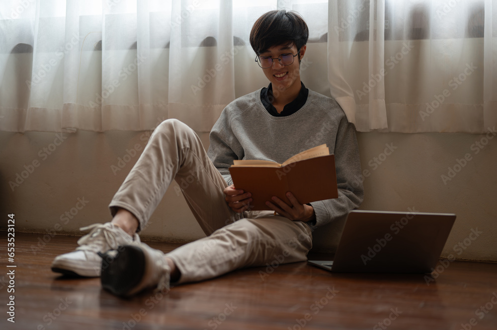 A happy young Asian man enjoys reading a book while sitting on the floor in a room. A happy young Asian man enjoys reading a book while sitting on the floor in a room.