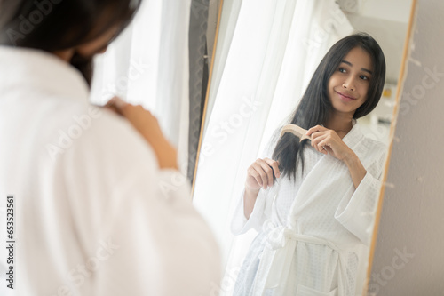 Beautiful lady in a bathrobe is combing her hair with a brush while standing in front of the mirror