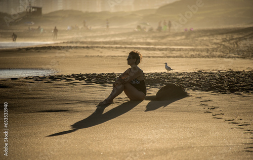 Mature woman on the beach sitting in front of the sea looks at the landscape on a golden afternoon, in the background the mist and buildings, in Punta del Este, Uruguay.. photo
