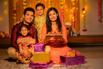 Portrait of happy young Indian family in traditional dress with lots of gifts around sitting on floor celebrating diwali festival at home.