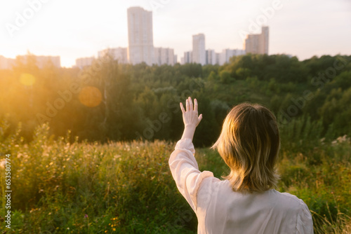 A girl with her hand raised stands on a mountain in the camera looking at the city in the distance at sunset