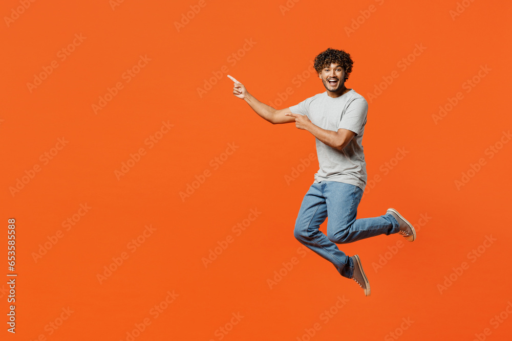 Full body young fun smiling happy Indian man he wears t-shirt casual clothes jump high look camera point index finger aside isolated on orange red color background studio portrait. Lifestyle concept.
