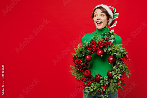 Young amazed woman wear green turtleneck Santa hat posing hold in hand decorative wreath look aside on area isolated on plain red background. Happy New Year 2024 celebration Christmas holiday concept. photo