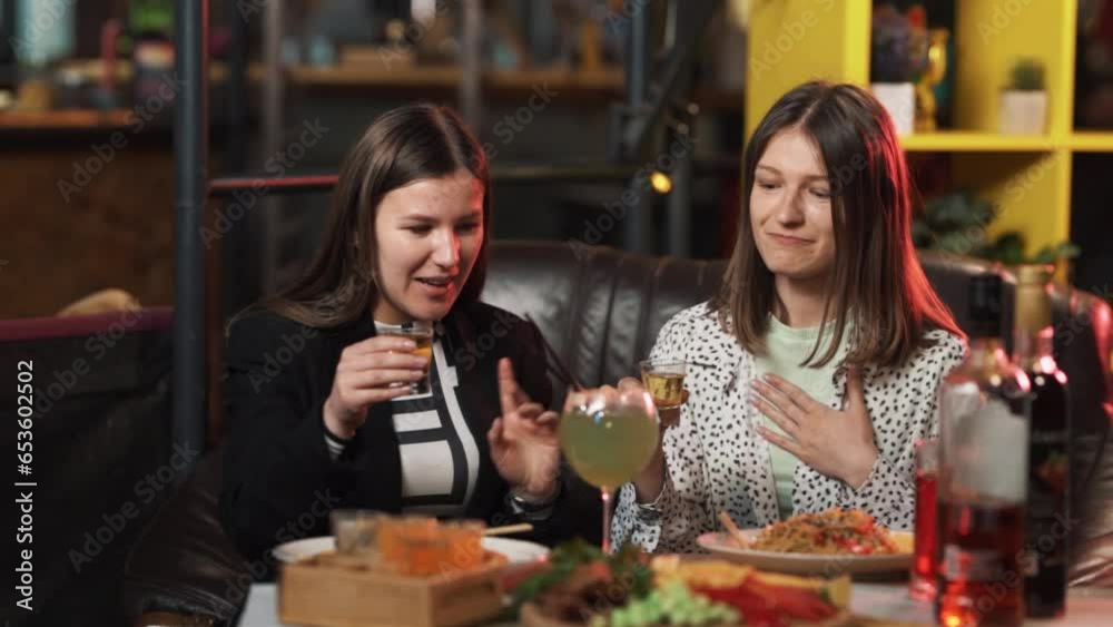 Two girls, sitting at a table in the bar, are drinking alcoholic shots Two girls, sitting at a table in the bar, are drinking alcoholic shots