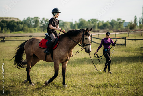 A teenage boy in a helmet learns horse riding in the summer, the instructor teaches the child equestrianism.