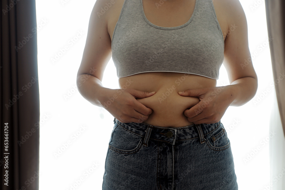 Close up of a belly with scar from c-section and abdominal fat. Women's health. A woman dressed up in sportswear demonstrating her imperfect body after a childbirth with nursery on the background. - obrazy, fototapety, plakaty 