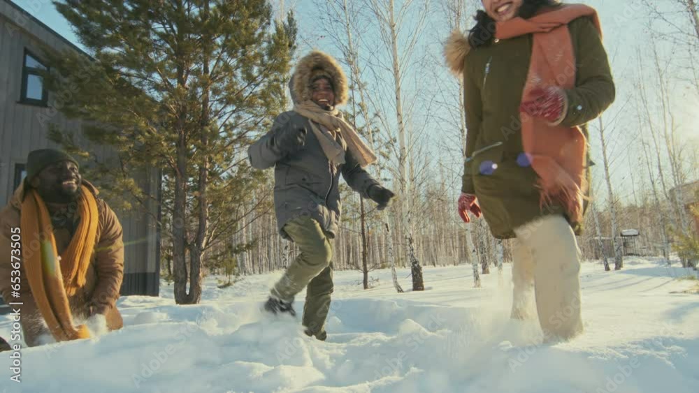 Handheld low angle shot of joyful interracial family with kid running around and playing with snow on sunny winter day