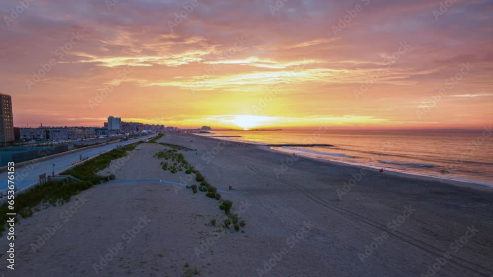 An aerial time lapse over Rockaway Beach, New York during a beautiful and cloudy sunrise. The drone camera starts high over the water, then truck left and boom down to the beach as the sun rises. An aerial time lapse over Rockaway Beach, New York during a beautiful and cloudy sunrise. The drone camera starts high over the water, then truck left and boom down to the beach as the sun rises.