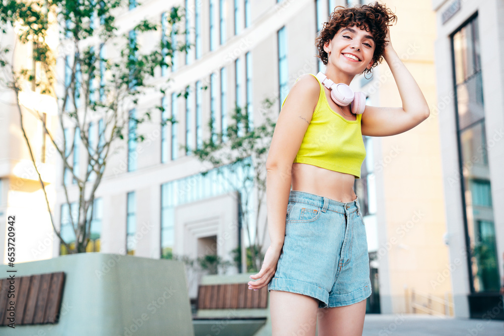 Young beautiful smiling hipster woman in trendy summer clothes. Carefree woman with curls hairstyle, posing in the street at sunny day. Positive model outdoors. Listens music at her earphones