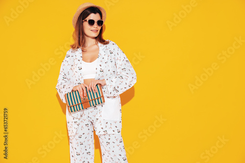 Young beautiful smiling female in trendy summer costume clothes. Carefree woman posing near yellow wall in studio. Positive model having fun indoors. Cheerful and happy. In hat. Holds clutch © halayalex