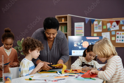 Portrait of kind Black woman helping group of little children doing crafts in preschool class, copy space