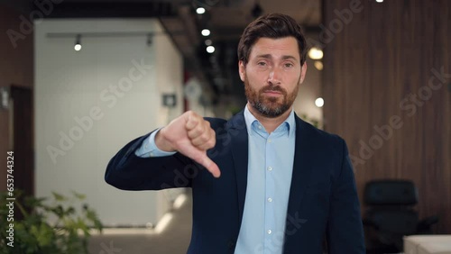 Bearded company worker looking unhappy and angry while showing rejection and negativity with thumbs down gesture. Young caucasian man wearing formal clothes standing over background of office area. photo