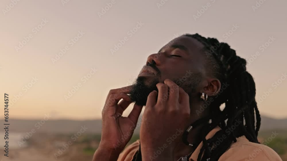 In a slow-motion, close-up shot, a black male closes his eyes, gently touches his face with his hands, and embraces a moment of self-awareness during a captivating sunset