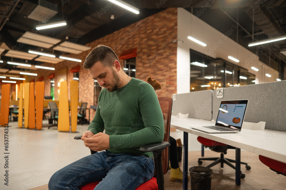 A bearded man uses a smartphone at his workplace in an open space office.