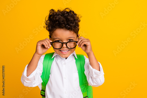 Portrait of toothy beaming smart boy with backpack wear stylish shirt touching glasses toothy smiling isolated on yellow color background