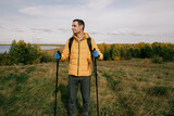 man with a backpack and gloves is walking with trekking poles on an autumn field against the background of the lake.An active lifestyle.Walking with trekking poles