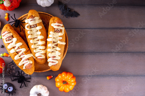 Funny mummy hot dogs for Halloween party or kids holiday brunch. Classic hot dogs with cheese eyes and cheesy mayo sauce, with Halloween decor, top view copy space