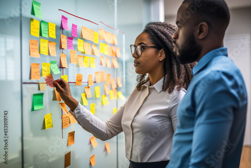 Young mixer race businesswoman manager explaining strategy ideas on sticky notes on glass wall to male African American colleague looking at strategy scrum presentation. Business project planning.