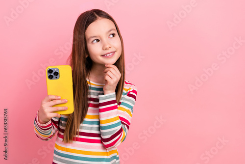 Photo of positive young small cute child girl touch her chin decide download new mobile games look mockup isolated on pink color background