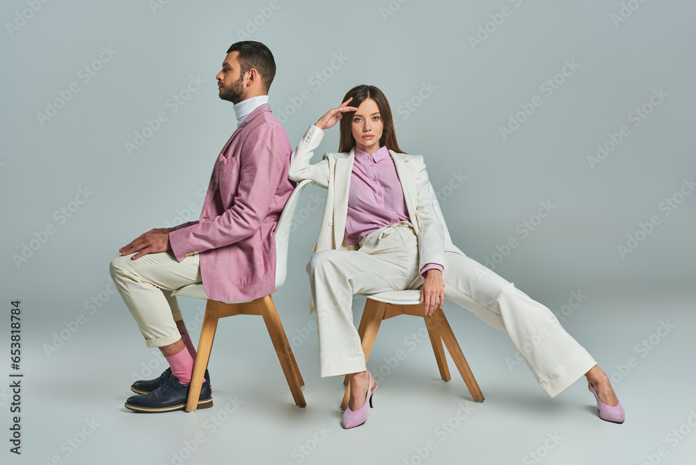 young woman in white suit sitting in armchair and looking at camera near man in lilac blazer on grey young woman in white suit sitting in armchair and looking at camera near man in lilac blazer on grey