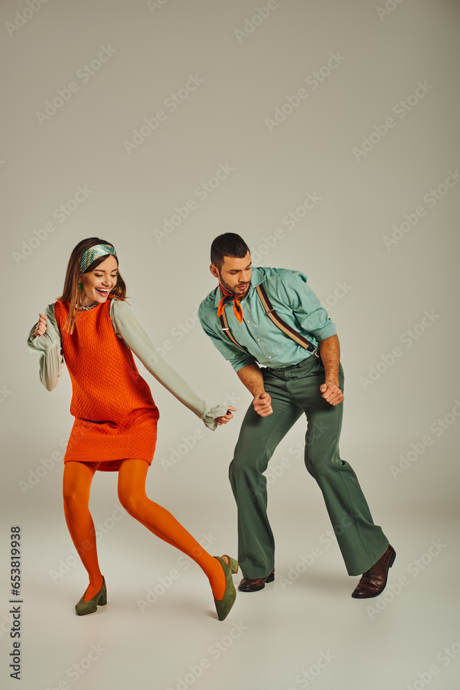 full length of young fashionable couple in colorful retro attire dancing on grey, fun and excitement