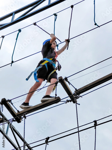 A teenage Caucasian boy in a rope amusement park crosses a suspension bridge