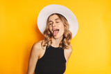 Portrait of naughty positive woman with wavy hair in white hat standing with closed eyes and demonstrating tongue, expressing disobedience disrespect, teasing grimace studio shot, yellow background.