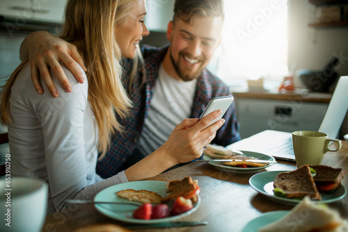 Young couple using a smartphone while having breakfast together in the morning in the kitchen at home