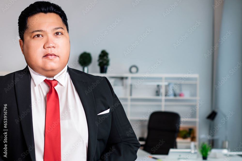 Male Asian plus size manager in black suit and red tie stands in the office Male Asian plus size manager in black suit and red tie stands in the office