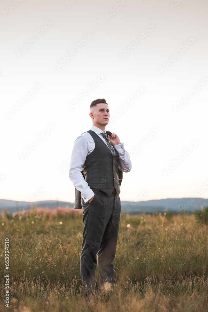 shot of a handsome young groom holding a jacket, photo in the middle of a field, the young man is stylishly dressed shot of a handsome young groom holding a jacket, photo in the middle of a field, the young man is stylishly dressed