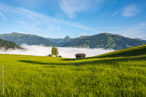 Alpine mountain landscape with green meadows, rural hayloft and Grosser Rettenstein Mountain on background. Sunny morning with weather inversion in the valley. Kitzbueheler Alps, Austria photo