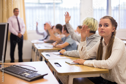 Diligent teenage students sitting with hands raised to answer during lesson in classroom