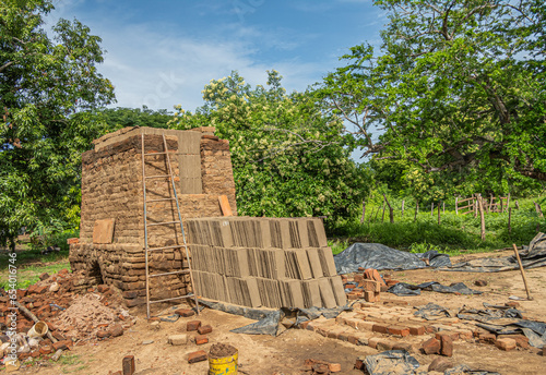 Zihuatanejo, Mexico - July 18, 2023: Historic Terracotta kiln set in its environment. Piles stones, bricks, dired panes on side. Ladder and green foliage as backdrop. Blue cloudscape  photo