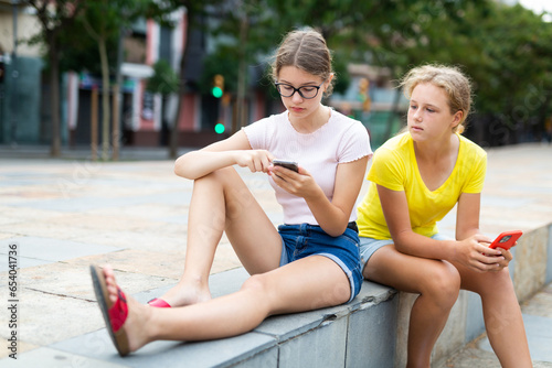 Two young girls sitting in park and using their smartphones.