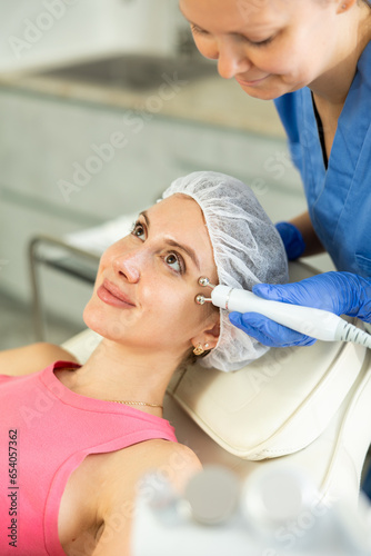 Adult woman cosmetologist performs facial microcurrent procedure with machine to young female client